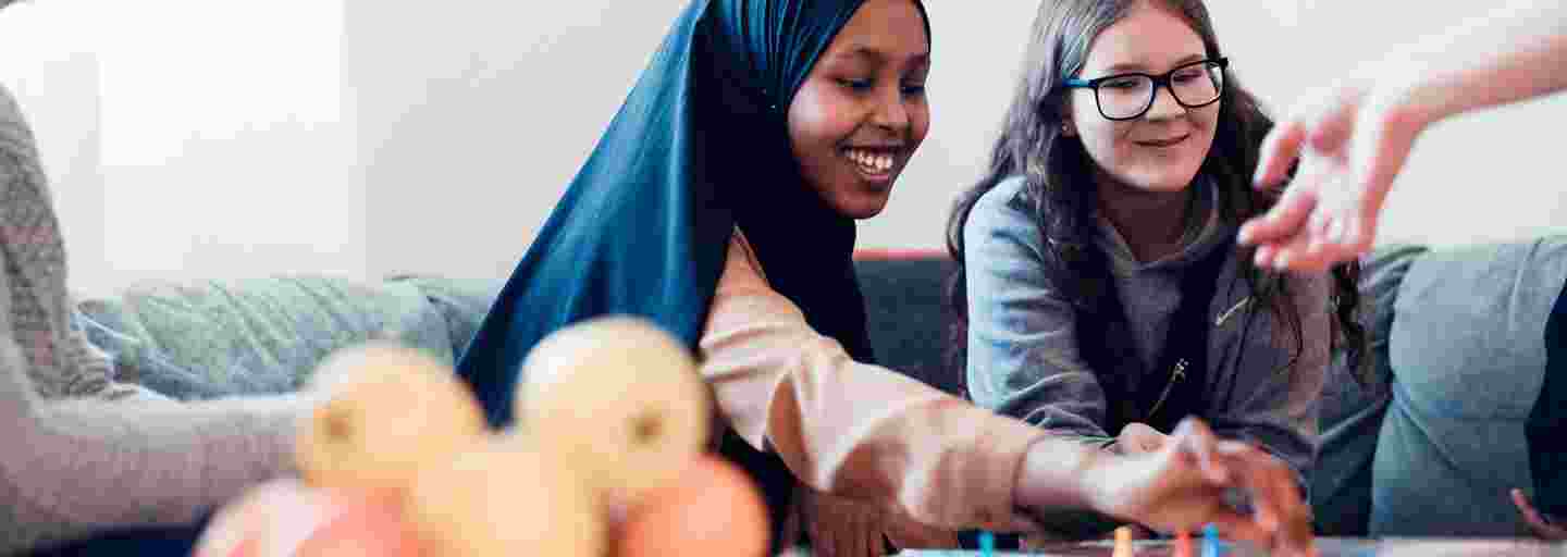 A smiling young person wearing a scarf playing a boardgame at a Youth Shelter with another young person.