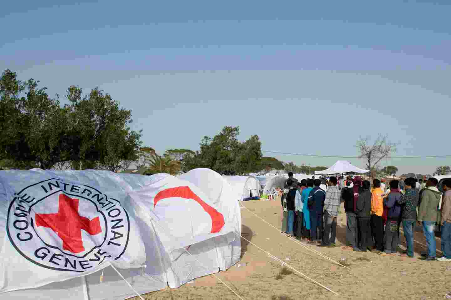 People queueing at a refugee camp among tents equipped with the Red Cross symbols.