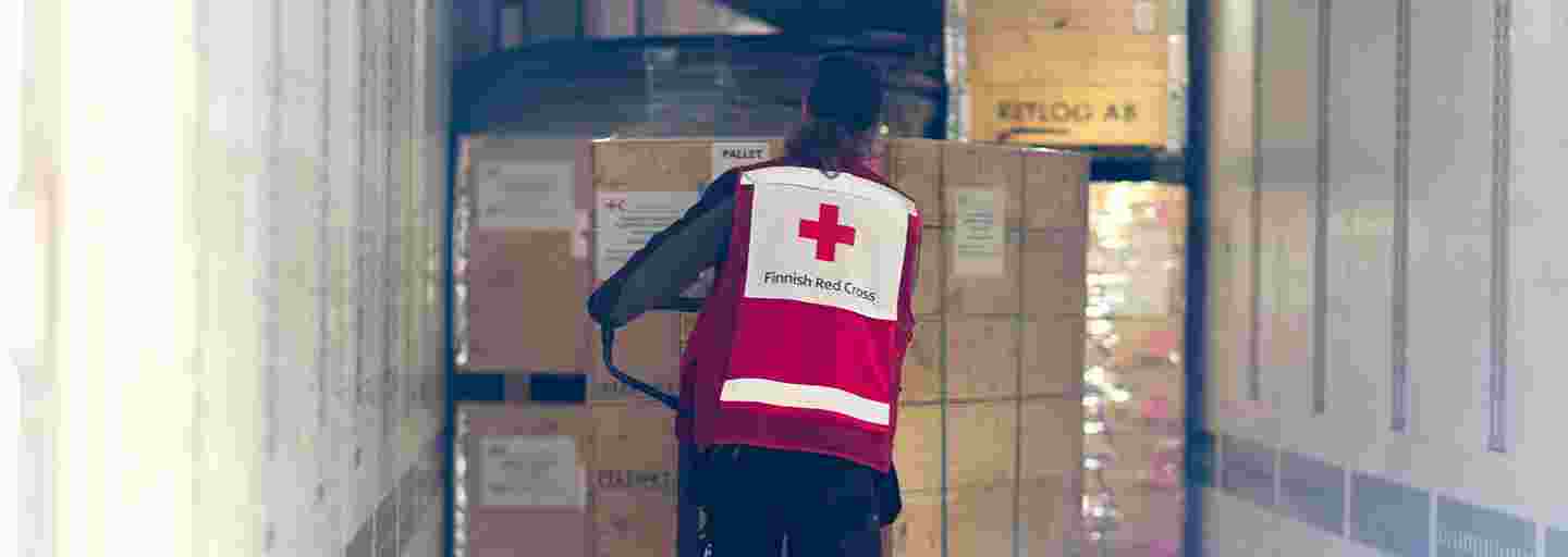 A Red Cross employee transporting a cart of large aid supply boxes down a storage hallway.