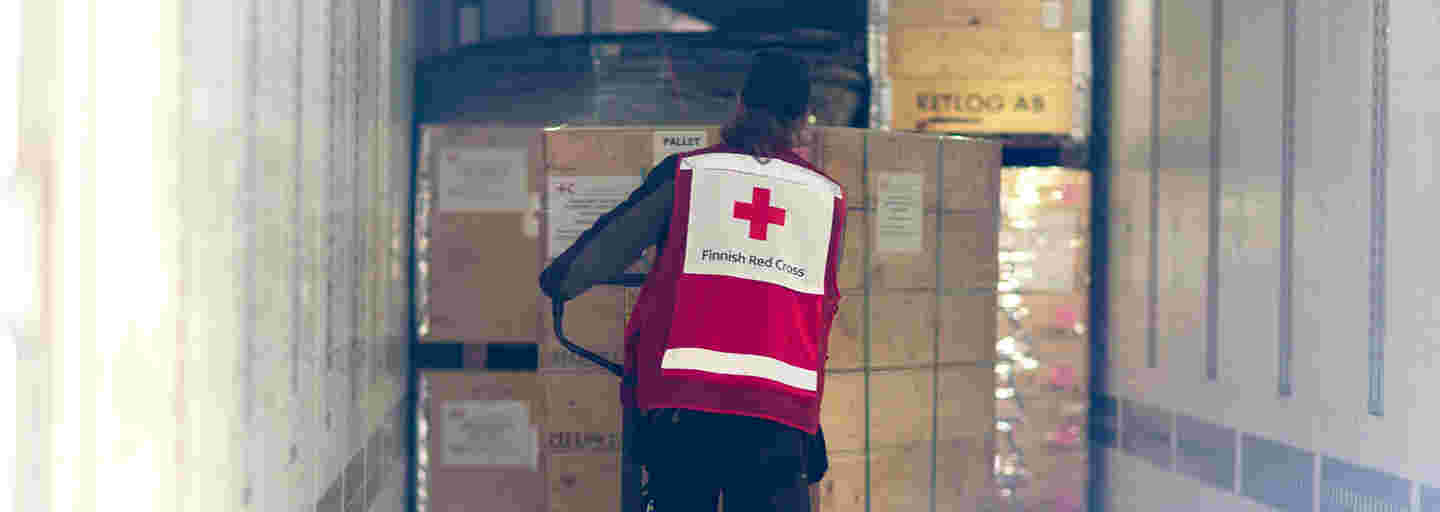 A Red Cross employee transporting a cart of large aid supply boxes down a storage hallway.