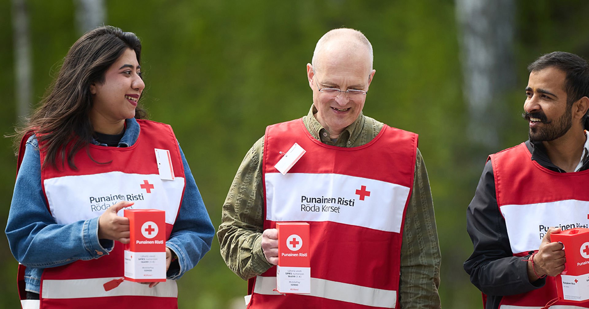 Three Finnish Red Cross Hunger Day fundraisers stand outdoors smiling and holding red collection boxes.